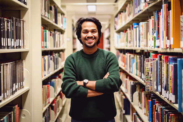 student kahirul standing among bookshelves in the library with his arms crossed and smiling