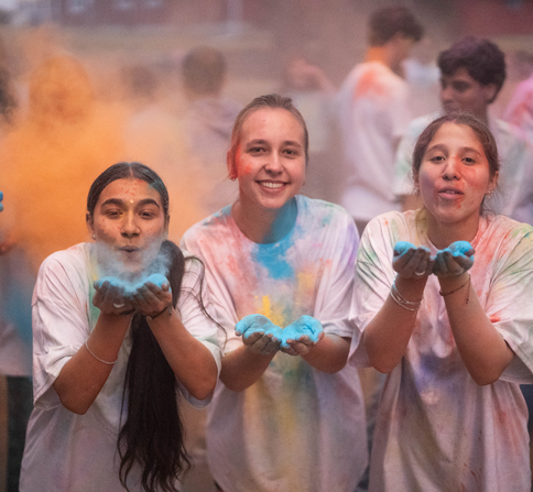 Image shows three girls, wearing white shirts during a Holi Celebration event (Hindu Festival) with a lot of different colored powder in the air and on their shirts.