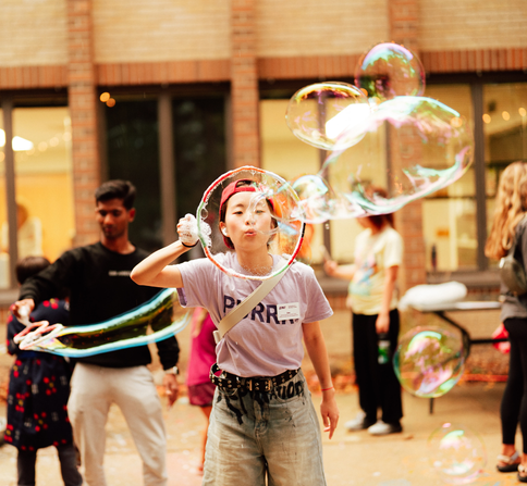 image shows a girls wearing a UNI shirt that says PURRRR, she is blowing bubble, with people in the background doing the same.